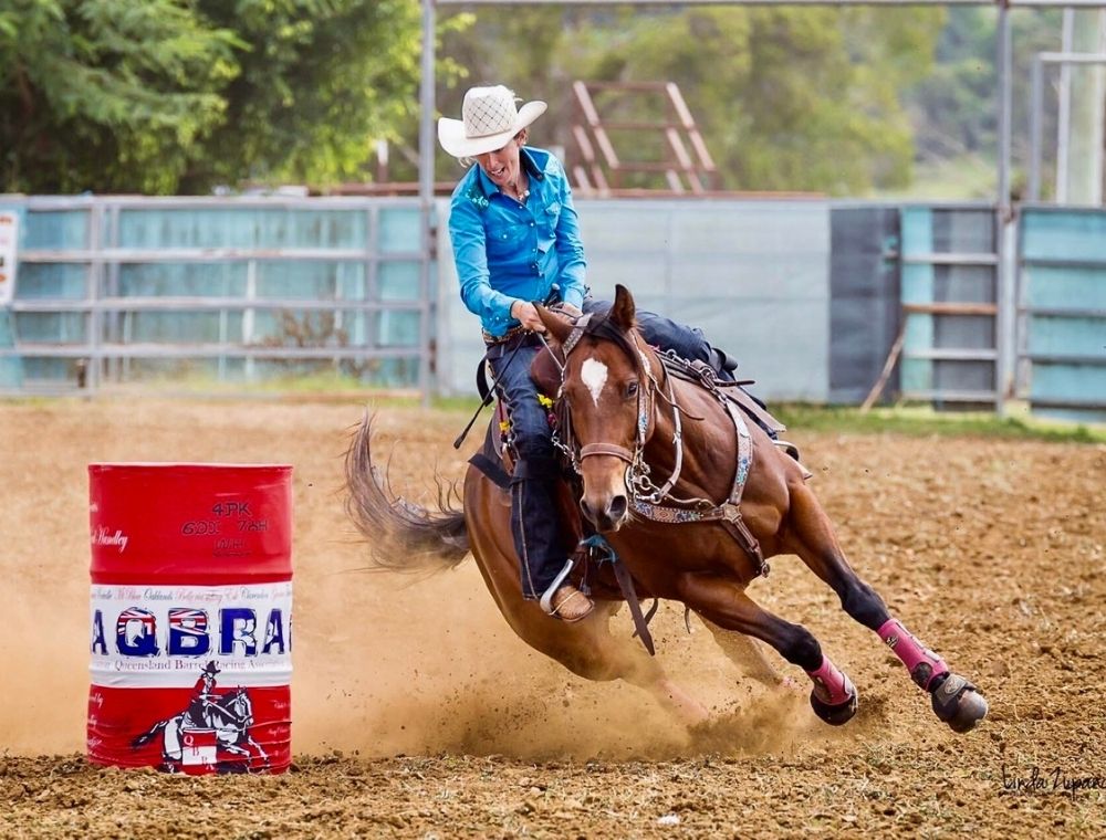 Photo of Kristy banks riding a brown horse. The horse is taking a sharp left turn around a red barrel. Kristy sits on top of the horse in a blue shirt, holding onto the reigns and smiling.