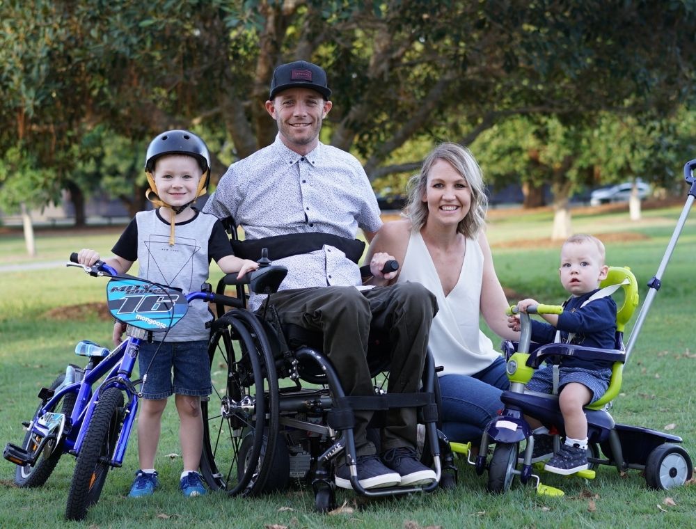 Photo of Dane sitting in his wheelchair at a park surrounded by his family. His wife is on the right of him in a white shirt. His eldest child stands on the left of him, holding a children's bike. On his right, next this himself and his wife, is their youngest child who is sitting on a bright green tricycle.