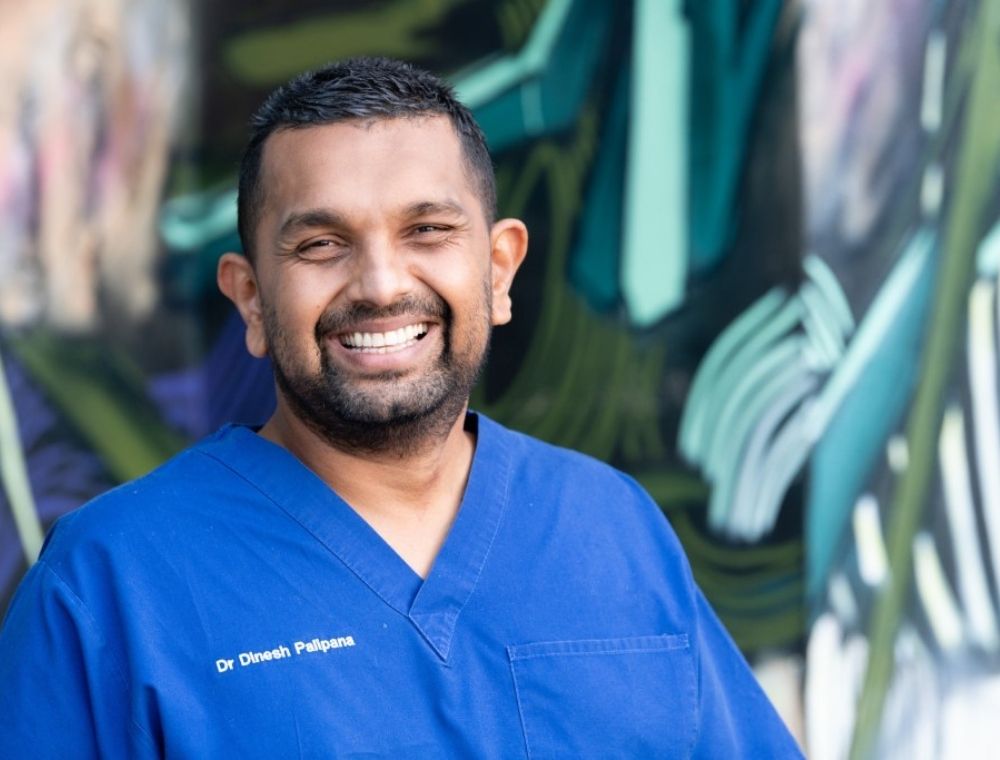 Photo of Dr Dinesh from the chest up. He is wearing blue medical scrubs with his name embroidered on the right-hand side. He has a big smile and is looking at the camera.  