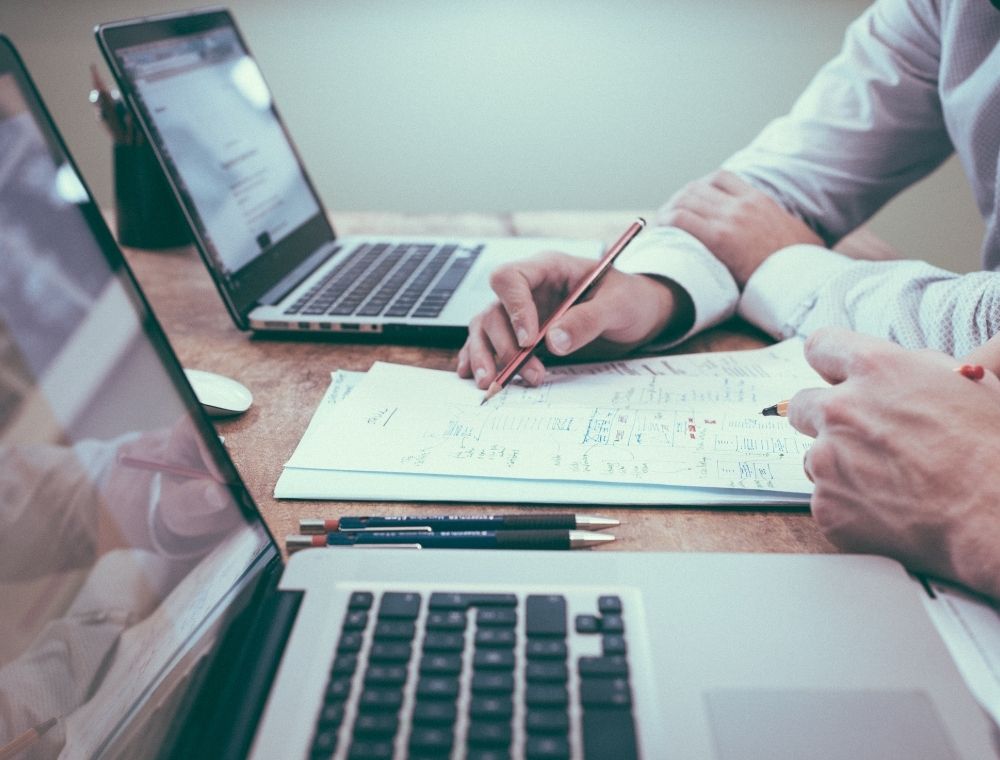 Photo of two hands holding a pencil and writing on a piece of paper. Next to the hands is two out of focus laptops. 