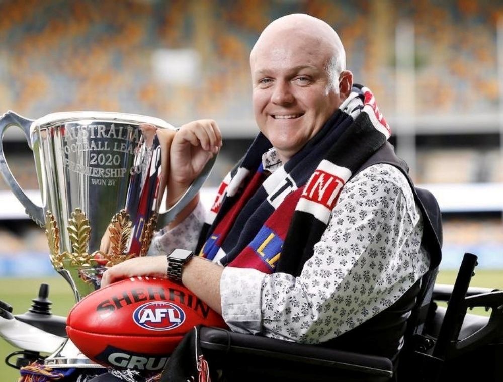 Tim McCallum sitting in his wheelchair smiling at the camera holding the 2020 Premiership Cup and red football.